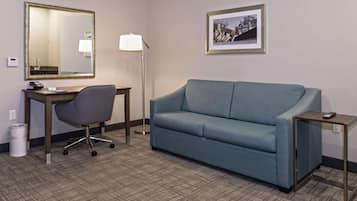 A hotel room with a desk, chair, sofa, lamp, framed picture, and a trash bin.