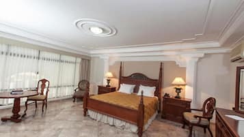 A hotel room with a large bed, two bedside tables with lamps, a wooden chair, a small table, and a large window with curtains.