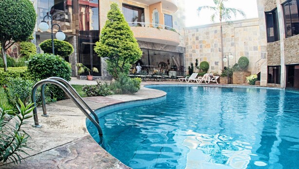 Ein Schwimmbecken mit Liegestühlen, umgeben von einem angelegten Garten und einem mehrstöckigen Gebäude mit Balkonen.