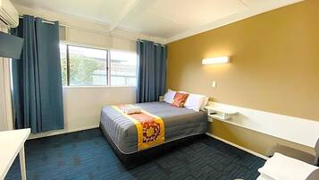 A hotel room with a bed, a desk, a chair, a window with curtains, and a wall-mounted light.