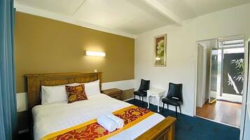A hotel room with a large bed, a bedside table, a chair, a small table, and a door leading to a balcony.