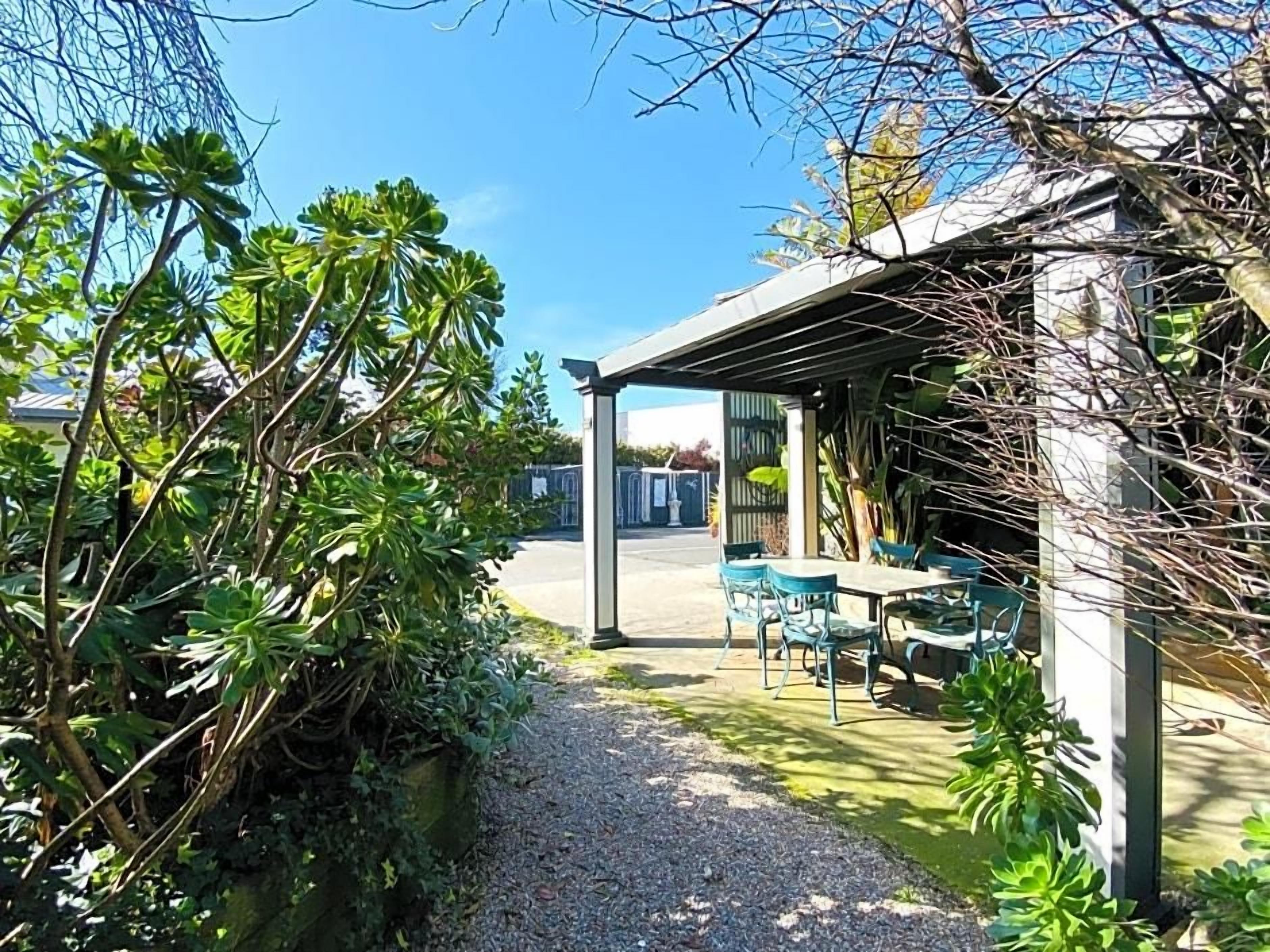 A covered outdoor seating area with tables and chairs, surrounded by greenery and a gravel path.