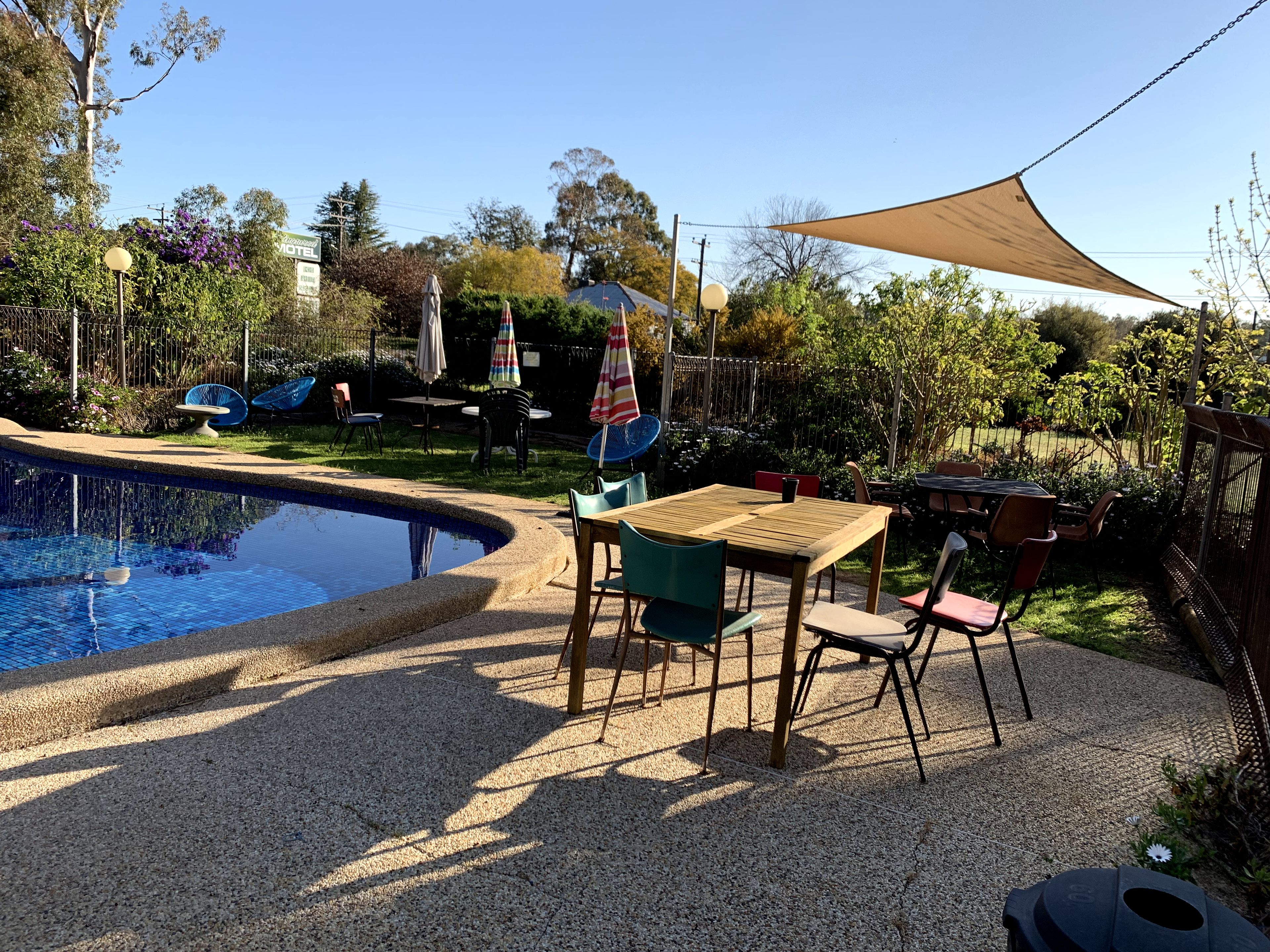 Zona junto a la piscina con mesa y sillas de madera, rodeada por una valla y árboles.