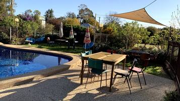 Zona junto a la piscina con mesa y sillas de madera, rodeada por una valla y árboles.
