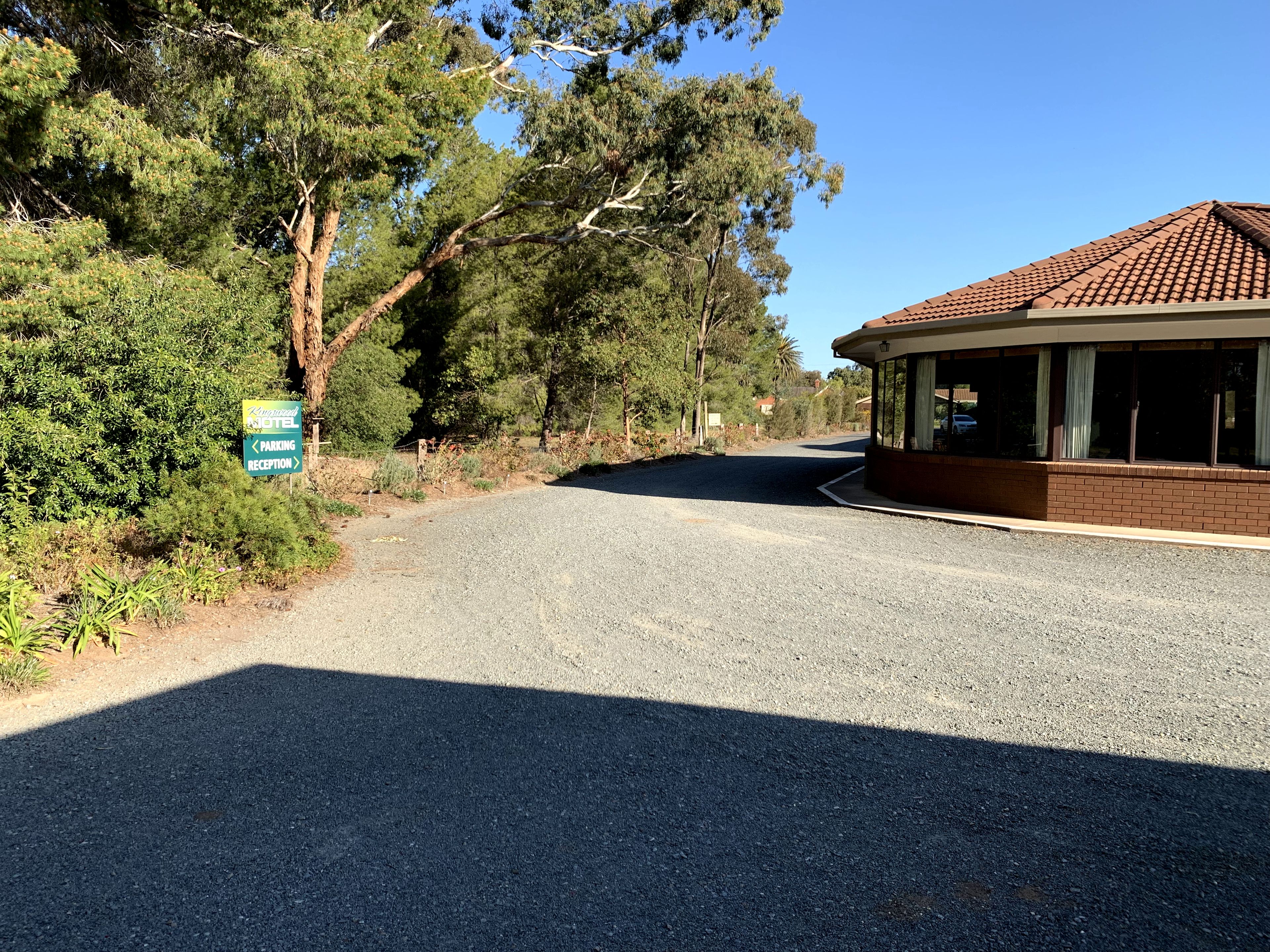 A gravel road leading to a building with a tiled roof, surrounded by trees and greenery. A sign indicates a parking area and reception.