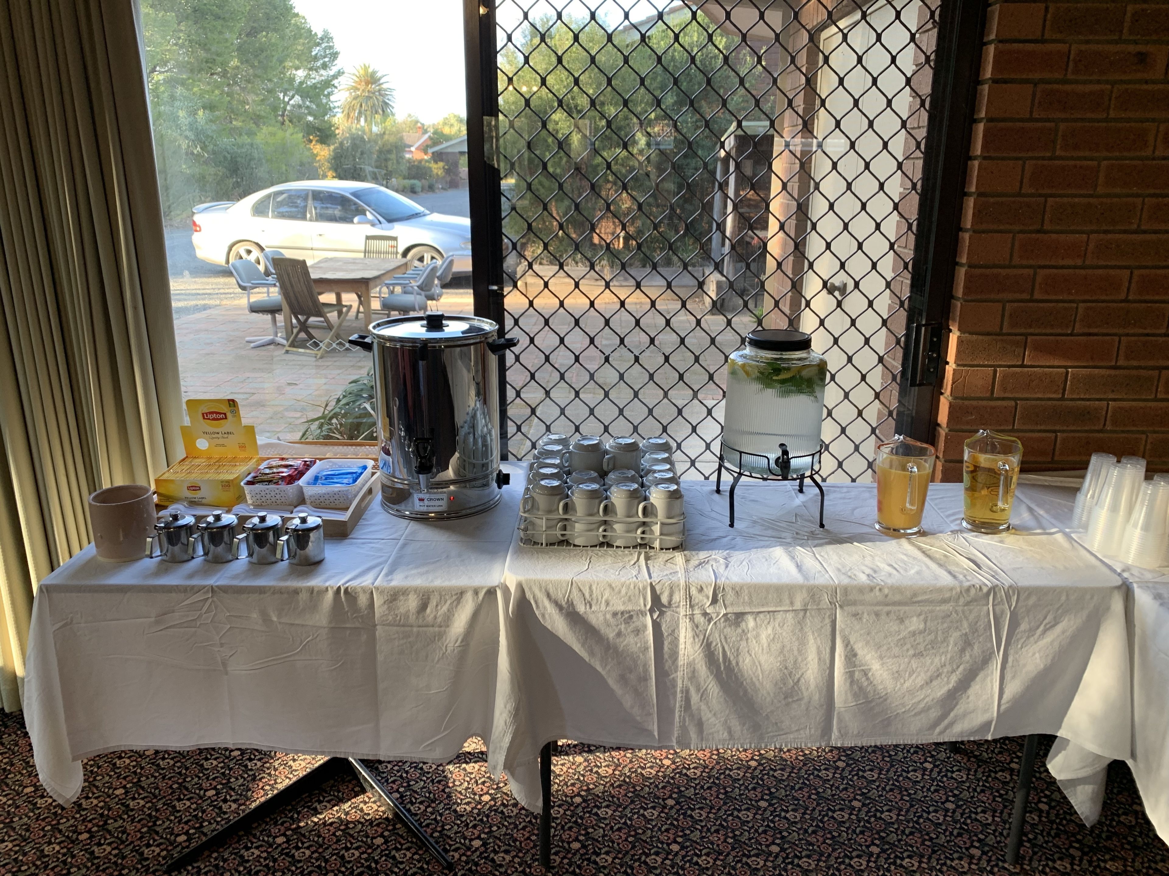 A table with a coffee station, including a coffee pot, cups, and a milk jug, set up near a window with a view of a parked car and trees.