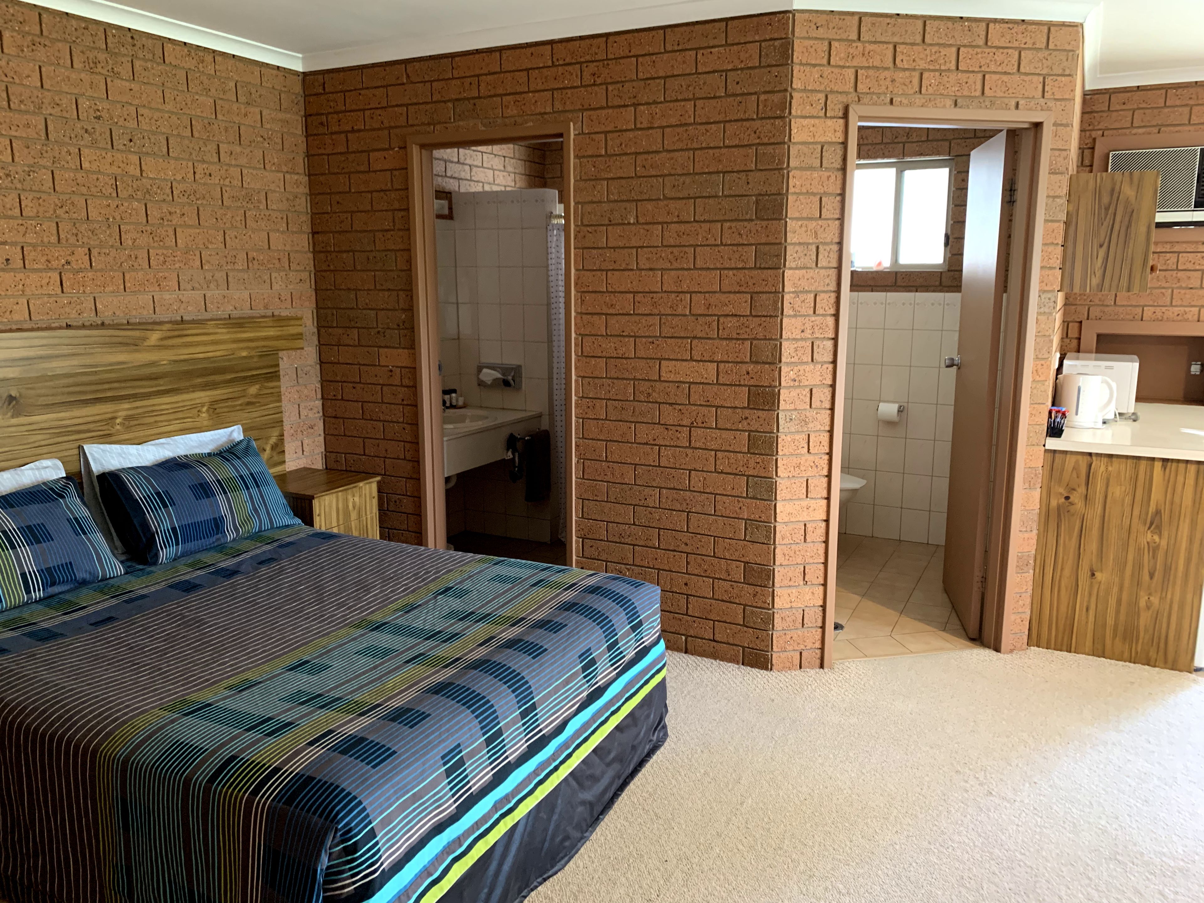 A bedroom with a brick wall, a wooden headboard, a bed with a striped bedspread, and a bathroom visible through an open door.