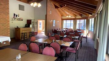 A spacious dining area with multiple tables and chairs, a kitchen area with a bar, and large windows providing natural light.