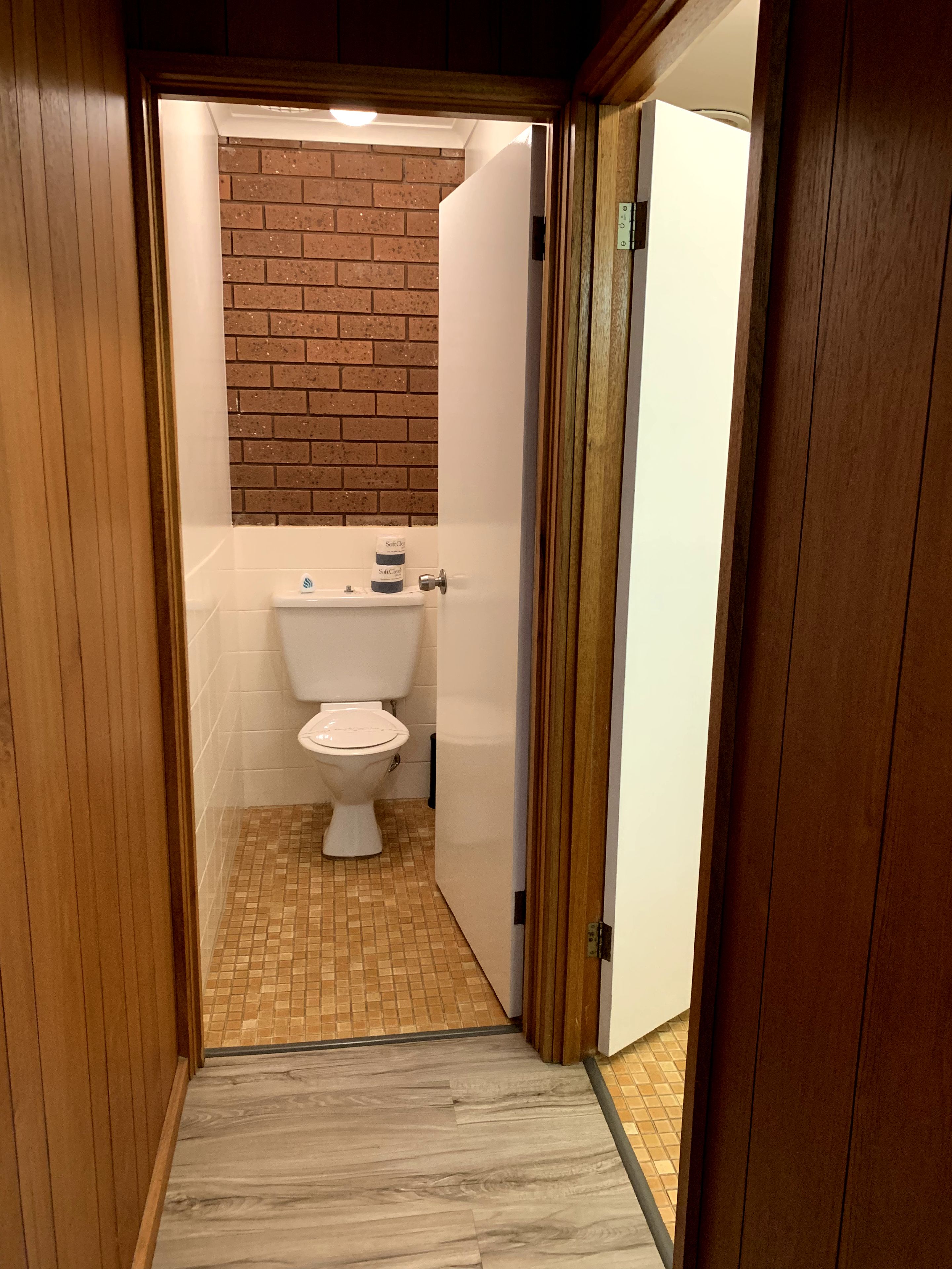 Un pasillo estrecho que da a un baño con un inodoro, un pequeño estante con artículos de aseo y una pared de ladrillo.