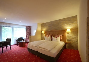 A hotel room with a double bed, two bedside tables with lamps, a desk with a chair, and a large window with curtains. - Flair Hotel Sonnenhof (BAIERSBRONN)