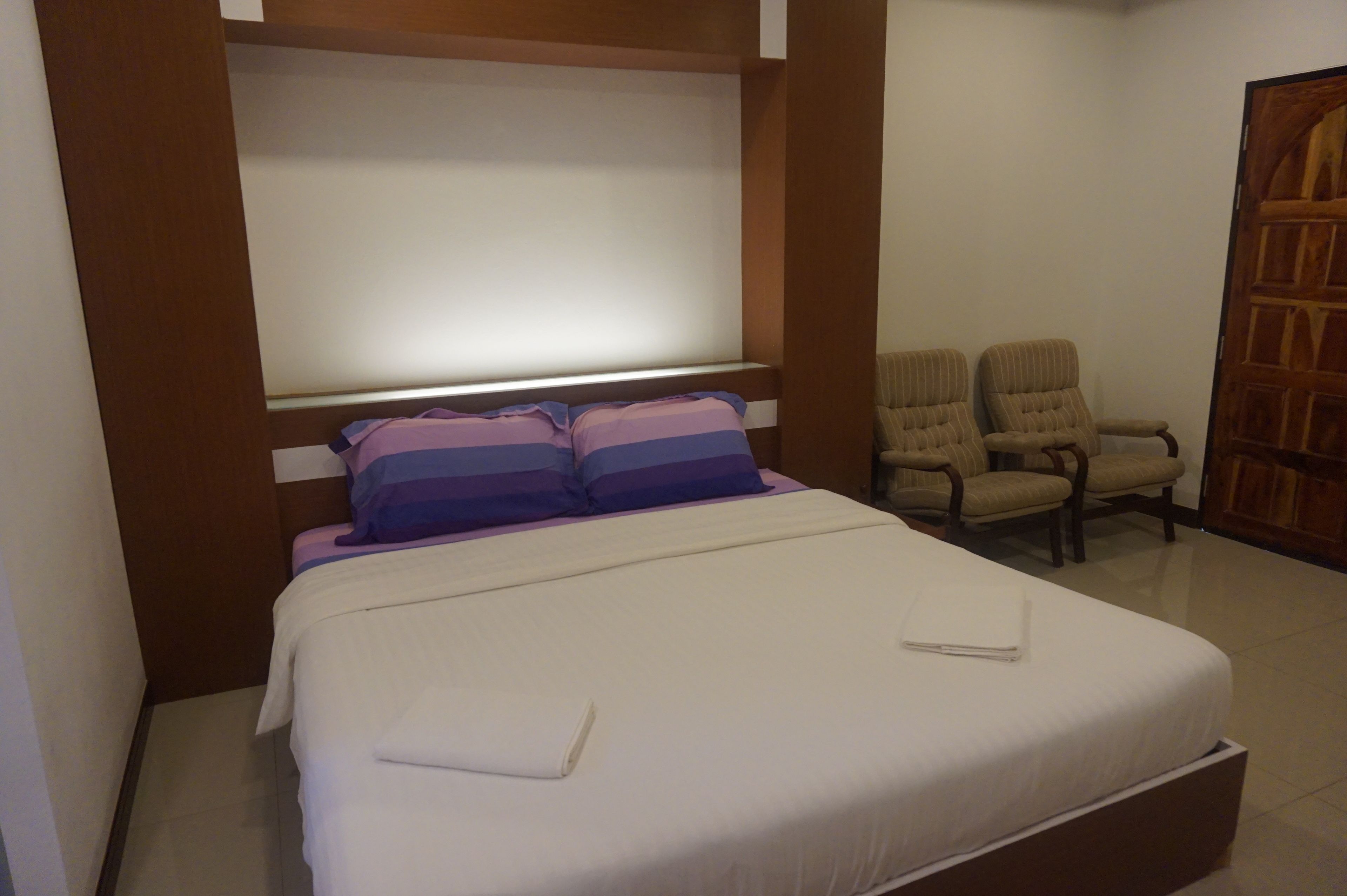 Un llit ben fet amb llençols de franfetina blanca i morada, dues cadires i un armari de fusta en una habitació d'hotel.