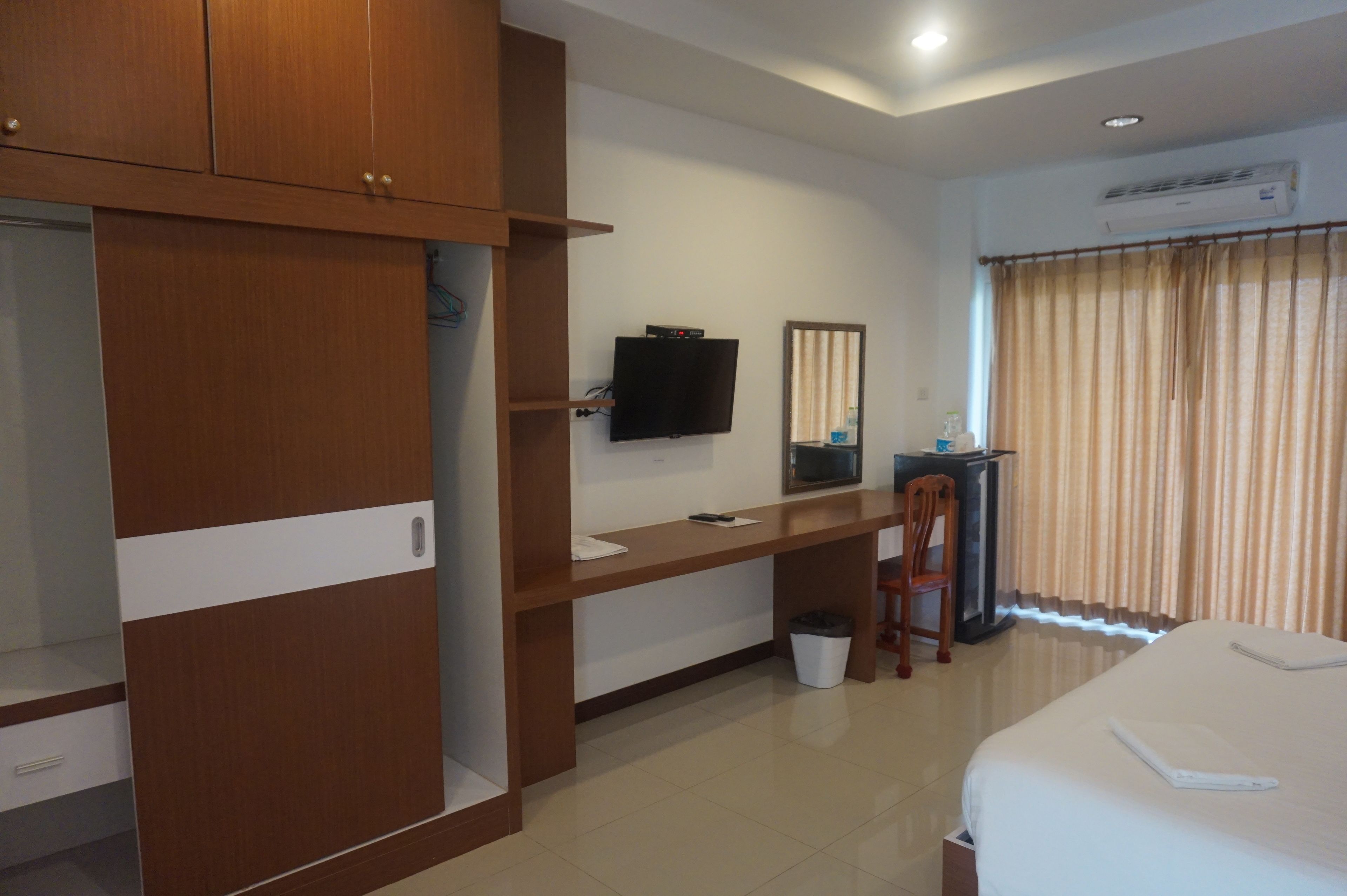 A hotel room with a wooden wardrobe, a desk with a flat-screen TV, a bed, and a window with curtains.