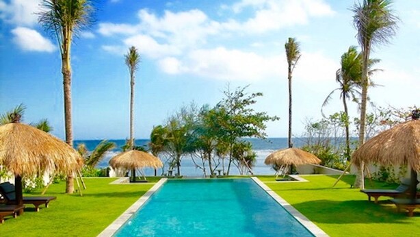 Una piscina rectangular con agua azul cristalina, rodeada de césped verde cuidado y sombrillas de paja.