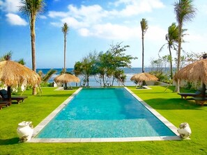 A rectangular swimming pool with a clear blue center, surrounded by well-maintained green grass and thatched umbrellas. - Villa Paradise Beach (Tabanan)