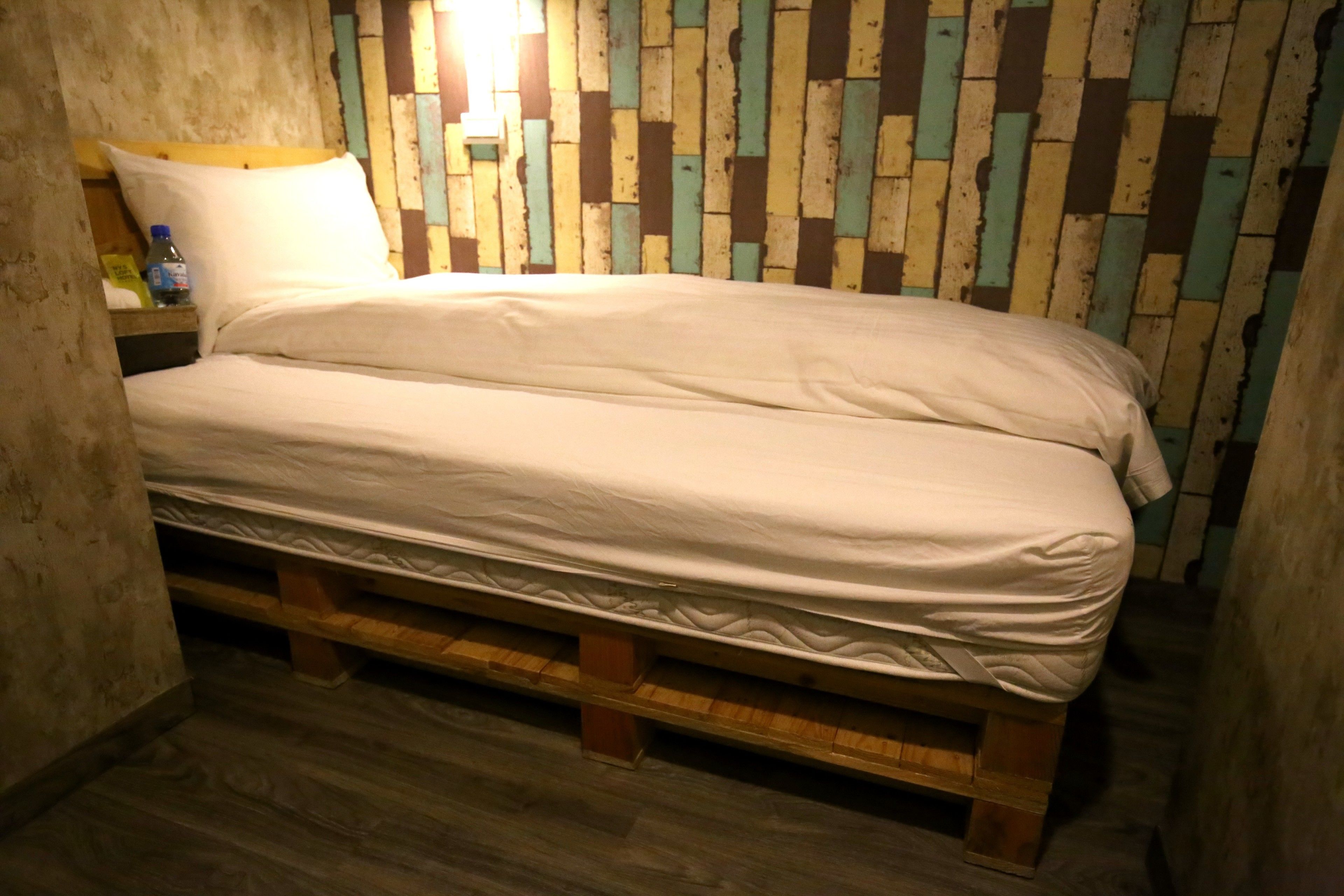 Un letto singolo con struttura in legno e materasso, appoggiato a un muro con pannelli di legno decorativi.