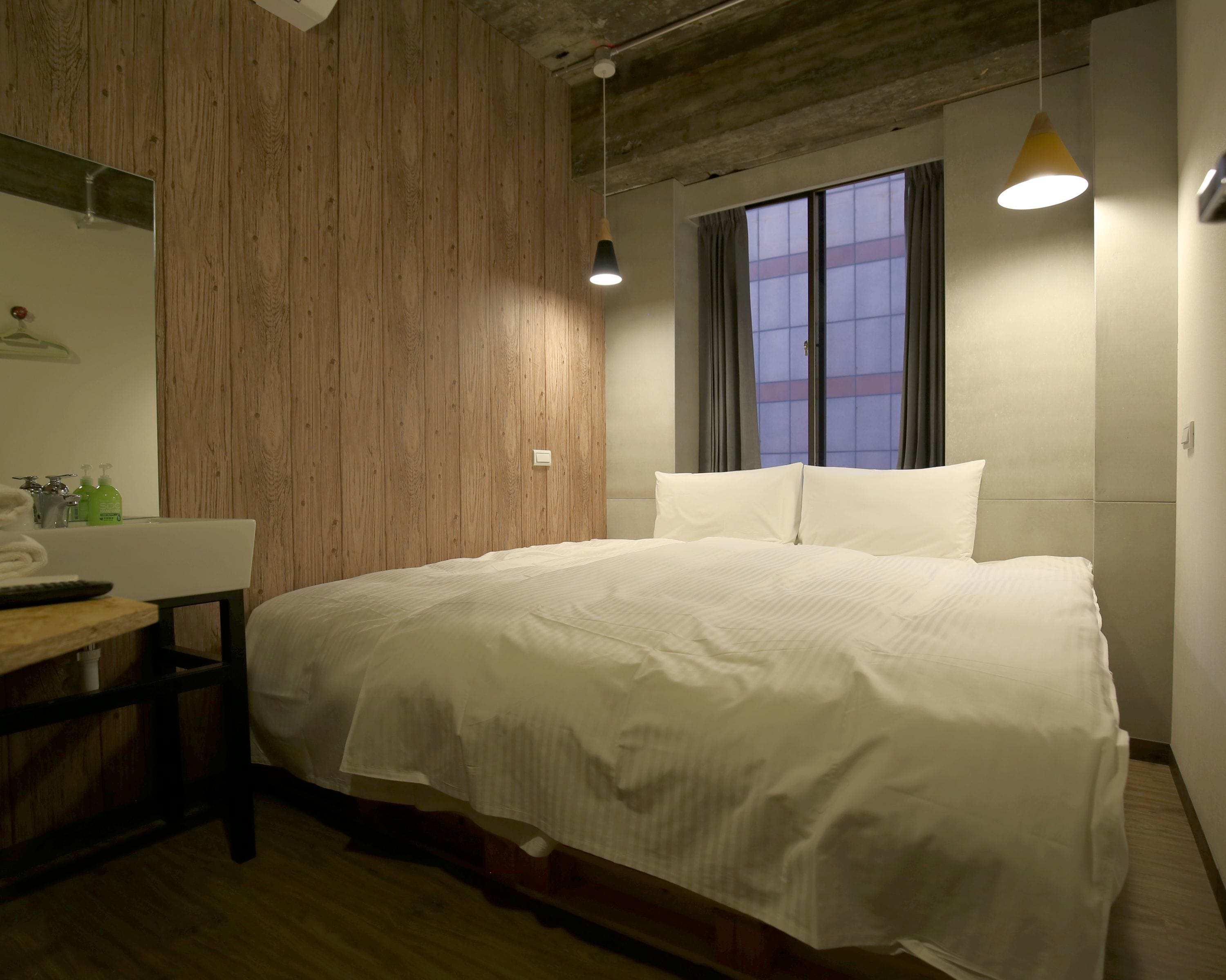 A bedroom with a wooden wall, a bed with white bedding, a bedside table, a mirror, and a window with curtains.