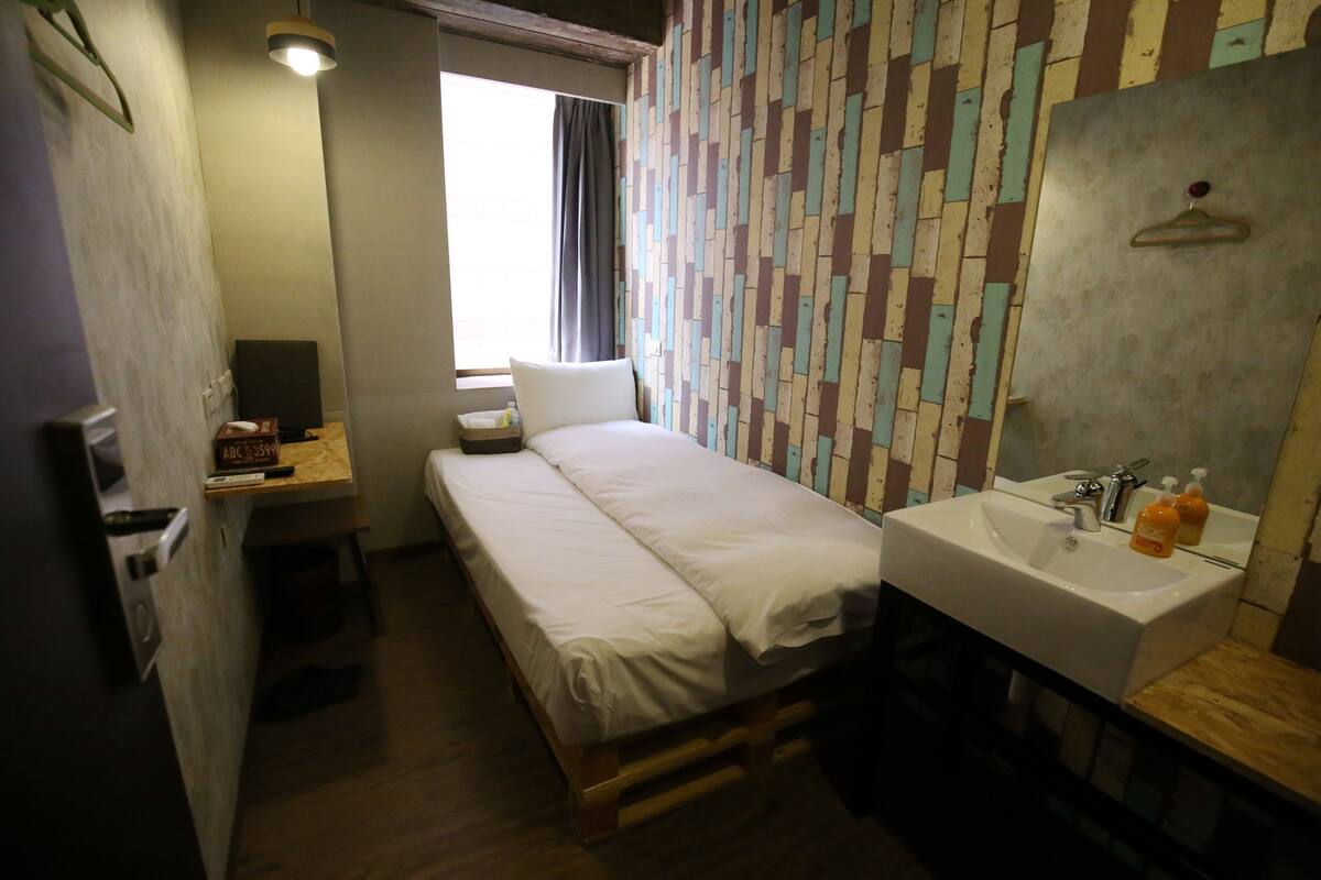 A small, compact room with a bed, a bedside table, a sink, and a mirror.