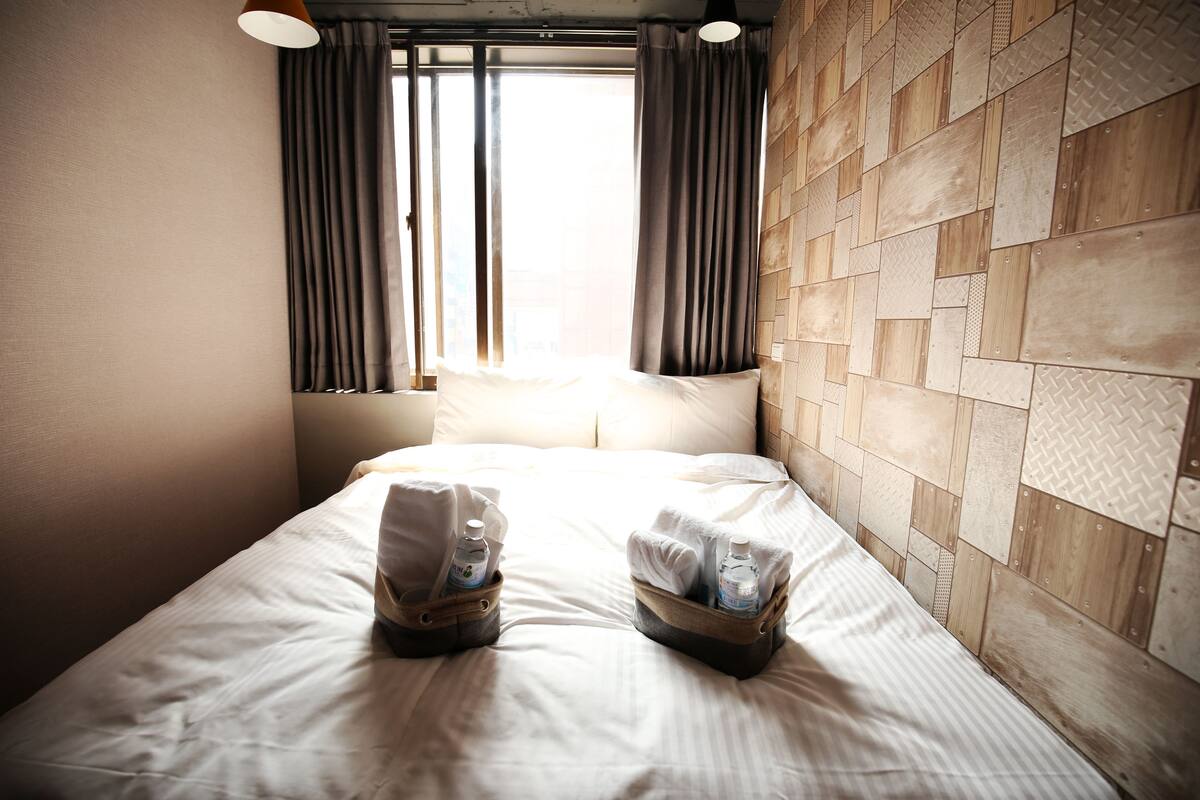 A hotel room with a bed, two bedside baskets with toiletries, a window with curtains, and a wooden wall panel.
