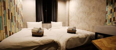 A double bed with white linens, a wooden nightstand with a bottle and a tissue box, and a wooden bench.
