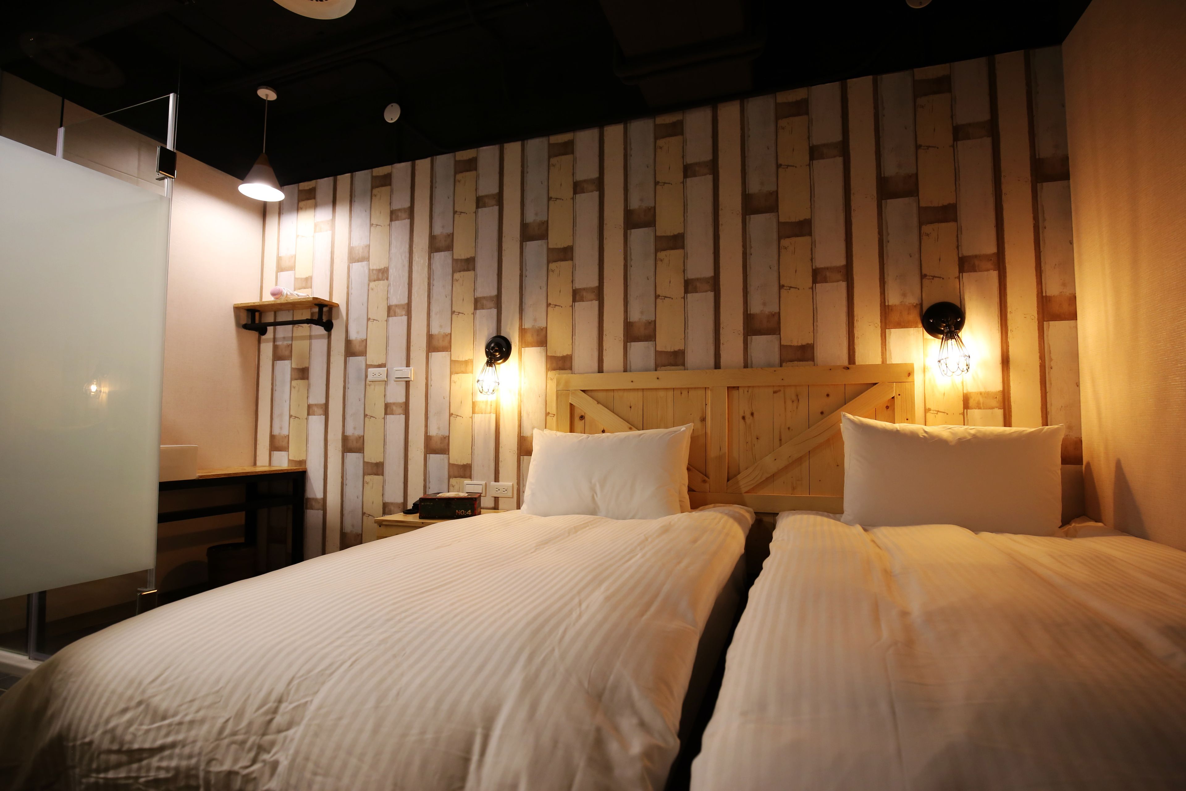 A hotel room with a wooden wall panel design, two beds with white bedding, a desk with a chair, and two wall-mounted lamps.