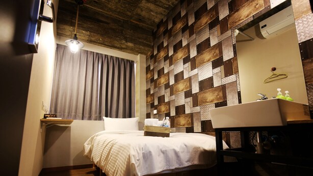 A small, cozy hotel room with a wooden ceiling, a bed with a white comforter, a bedside table with a lamp, a mirror, and a small shelf with toiletries.