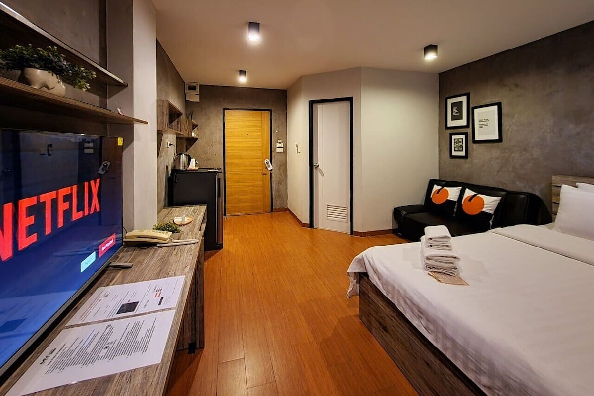 A modern hotel room with a wooden floor, a bed with white linens, a black sofa with orange pillows, a desk with a laptop, and a TV displaying the Netflix logo.