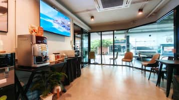 A modern coffee shop interior with a large TV screen, coffee machines, seating area, and a view of the street outside.