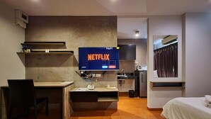 Ein modernes Hotelzimmer mit Schreibtisch aus Holz, einem Bett, einem Fernseher, der 'NETFLIX' anzeigt, und einer Küchenzeile.