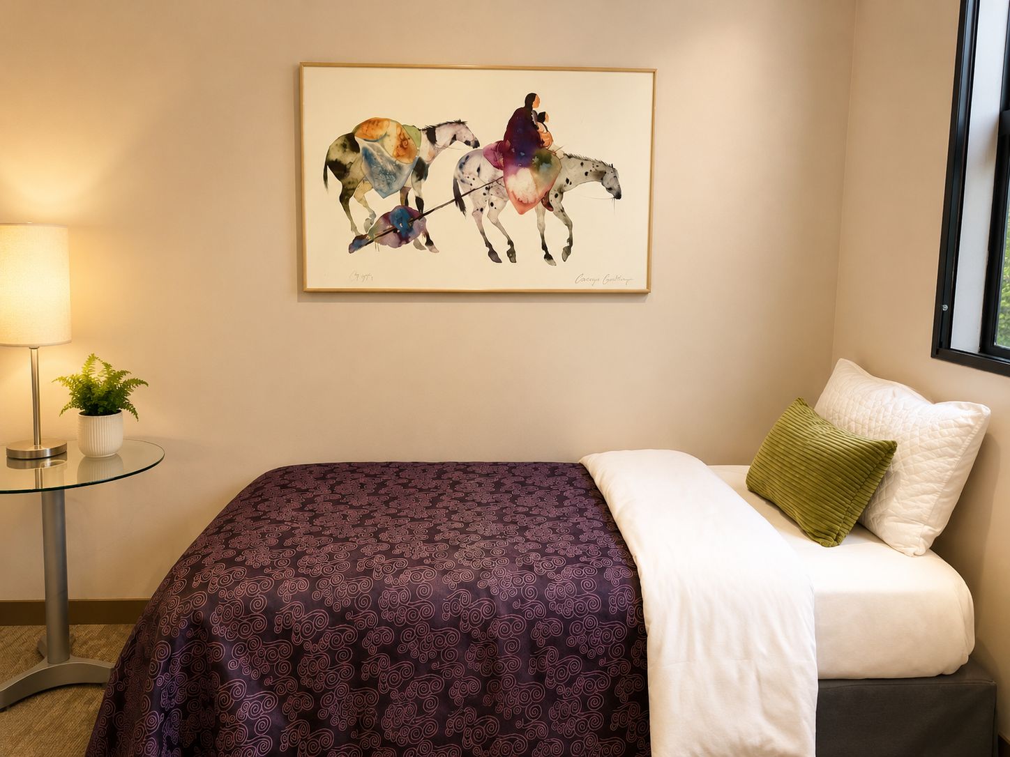 A hotel room with a bed, a framed artwork, a lamp, and a small table with a plant.
