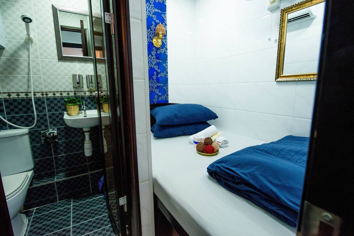 A bathroom with a shower, sink, and toilet. There is a bed with blue bedding and a decorative wall hanging.