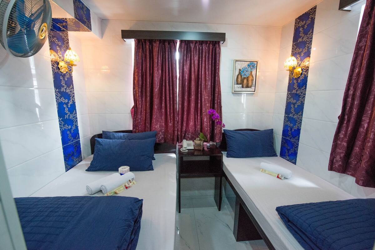 A hotel room with two beds, a small table, a fan, and a painting on the wall.