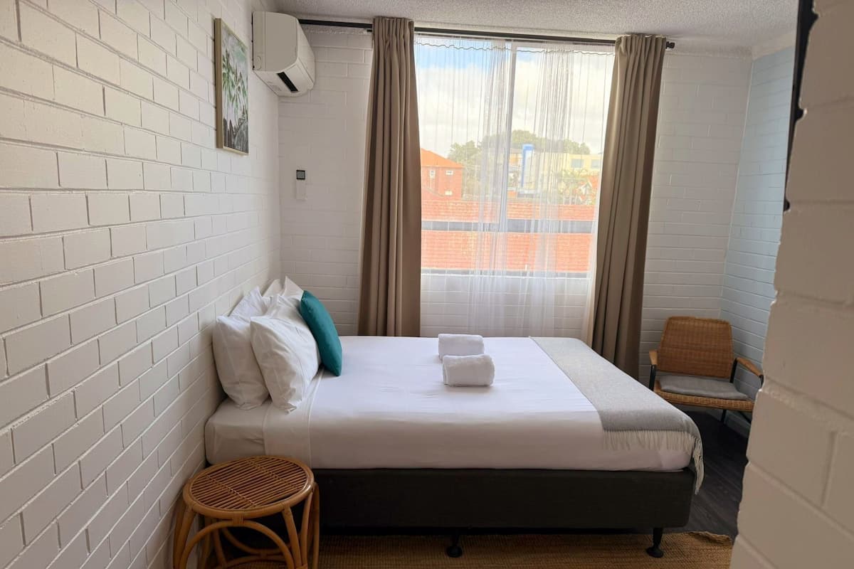 A neatly arranged hotel room with a bed, a small wicker stool, a chair, and a window with curtains.