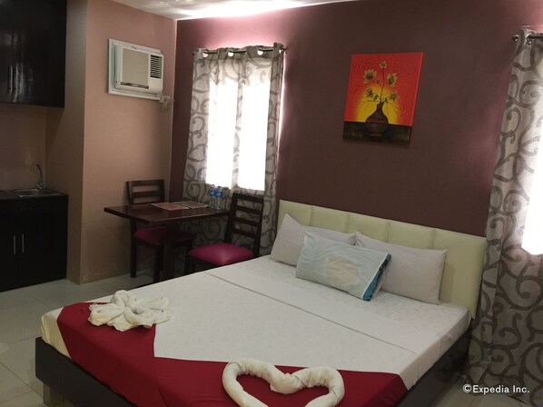 A hotel room with a bed, a desk with two chairs, a window with curtains, an air conditioning unit, and a painting on the wall. - Staylite Park B&B (Tagbilaran)