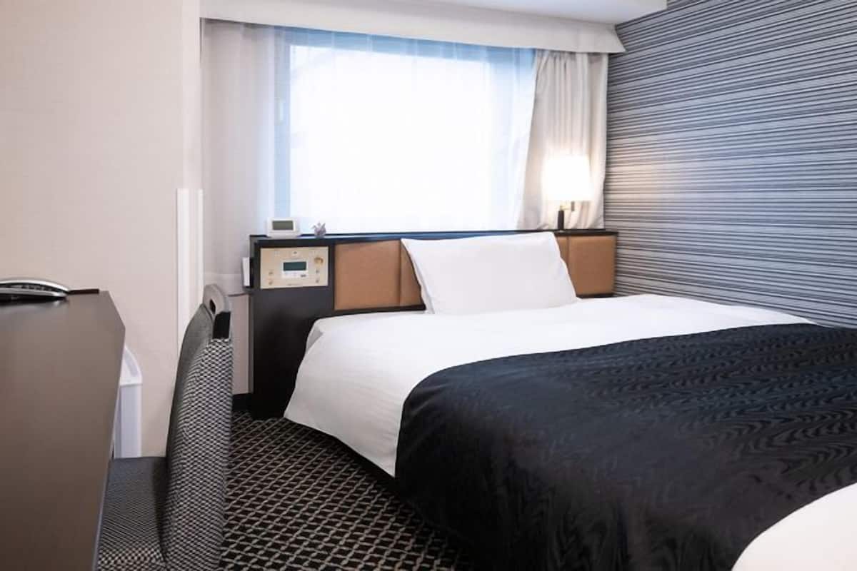 Habitación de hotel con cama, mesita de noche, lámpara, ventana con cortinas y pared con textura.