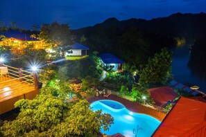 A resort with a swimming pool surrounded by lush greenery and multiple buildings with illuminated roofs. - Star Hill Riverkwai Resort (Sai Yok)