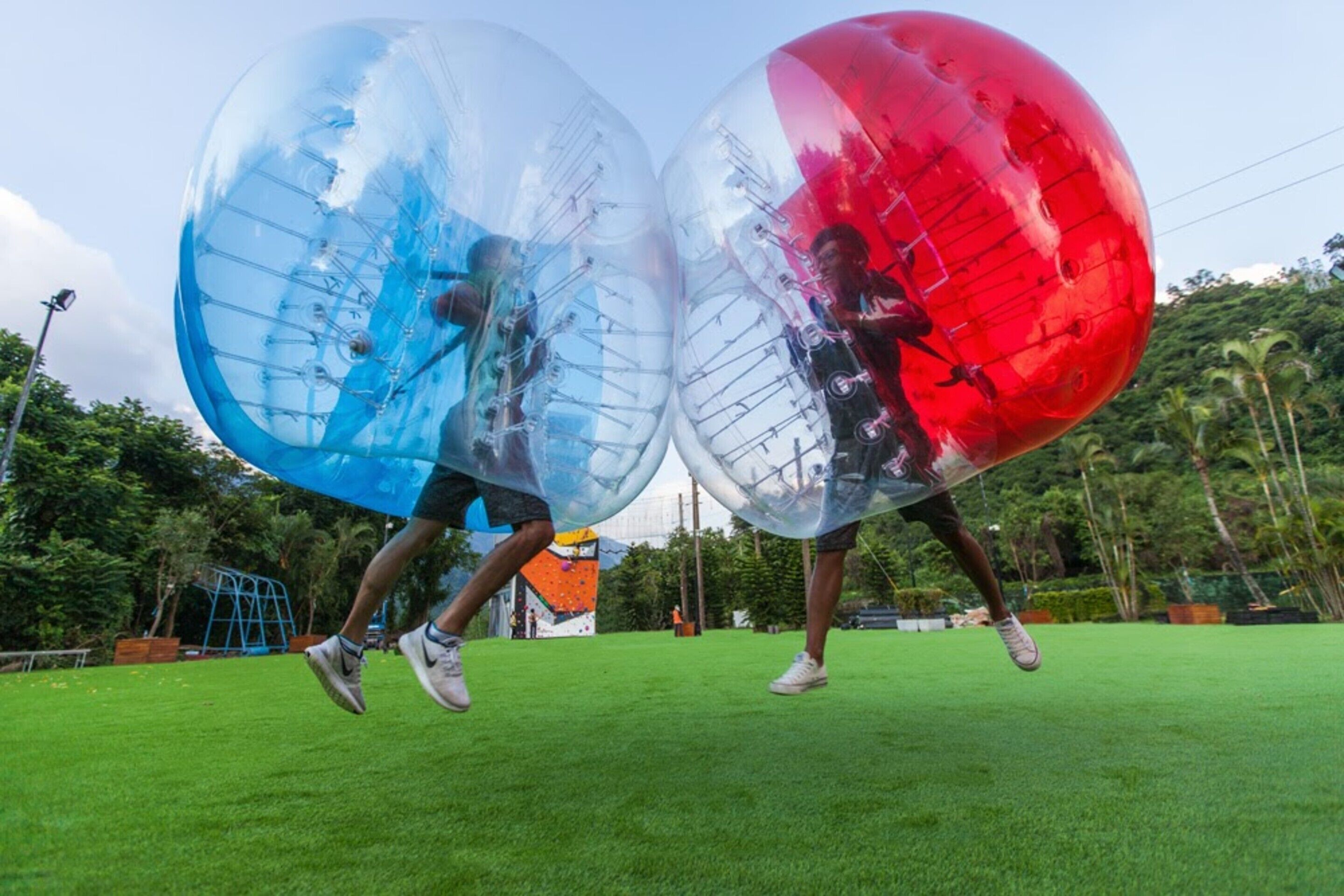 Dos personas dentro de esferas transparentes en un campo de hierba, con árboles y un parque infantil al fondo.