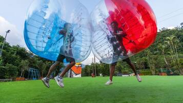 Dos personas dentro de esferas transparentes en un campo de hierba, con árboles y un parque infantil al fondo.