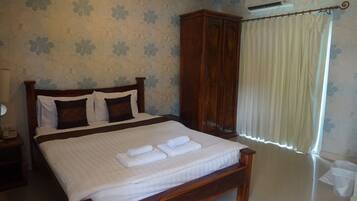 A neatly made bed with white linens and two folded towels, a wooden wardrobe, and a window with white curtains.