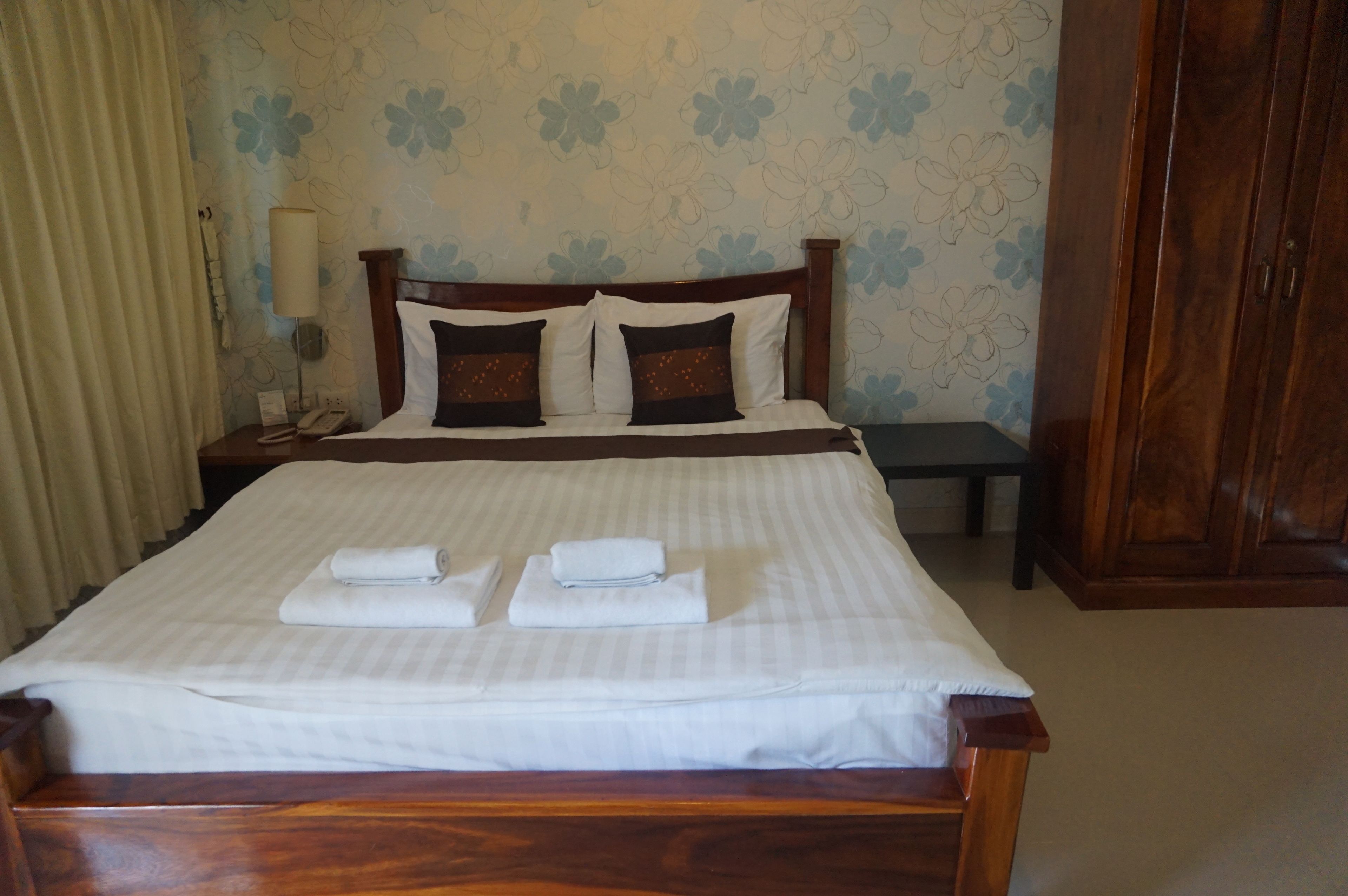A neatly made bed with white linens and two folded towels, a wooden headboard, a bedside table with a lamp, and a floral-patterned wall.