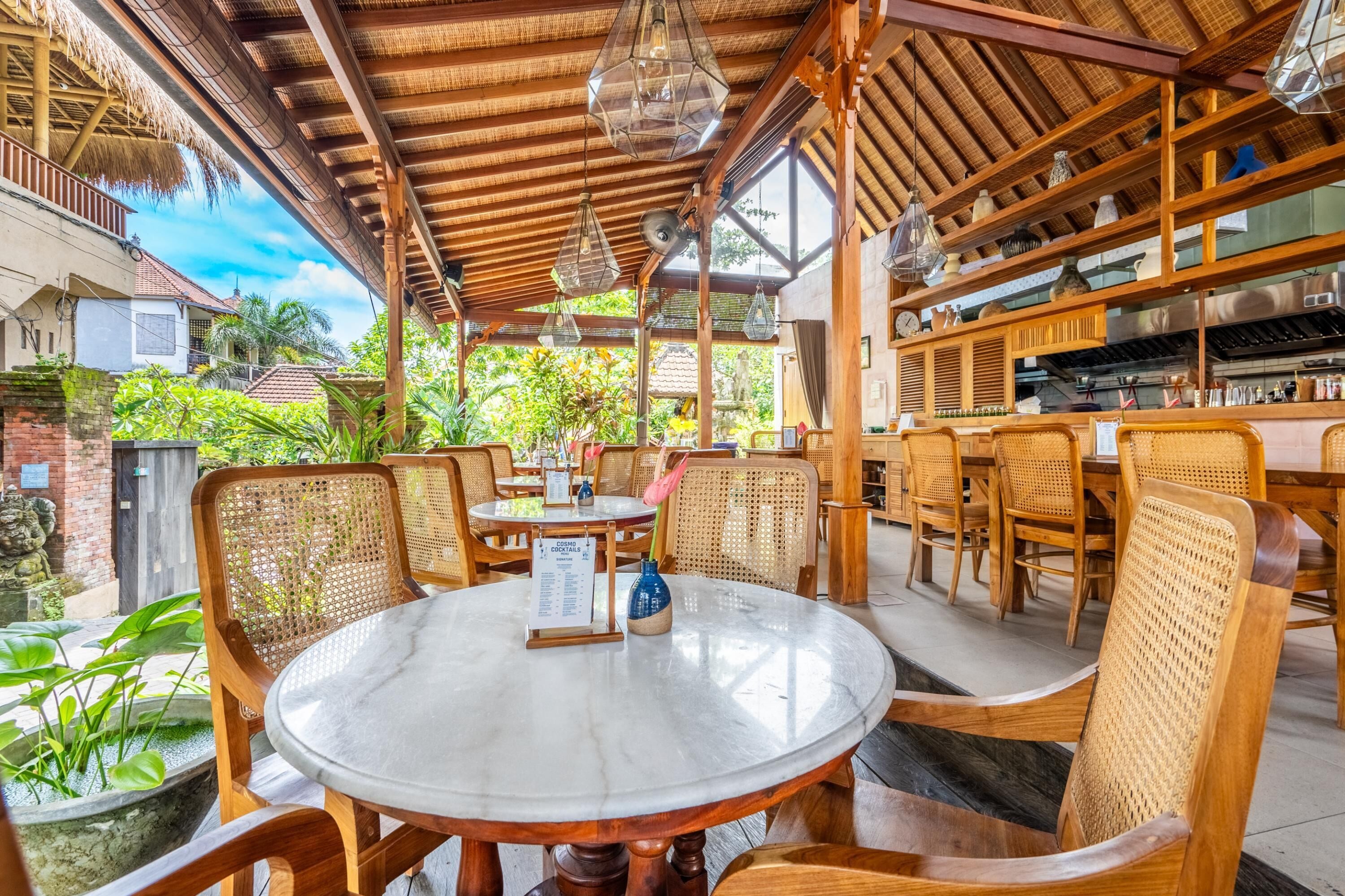 A restaurant with round tables and wicker chairs, a menu stand, and a kitchen area visible through the open sides.
