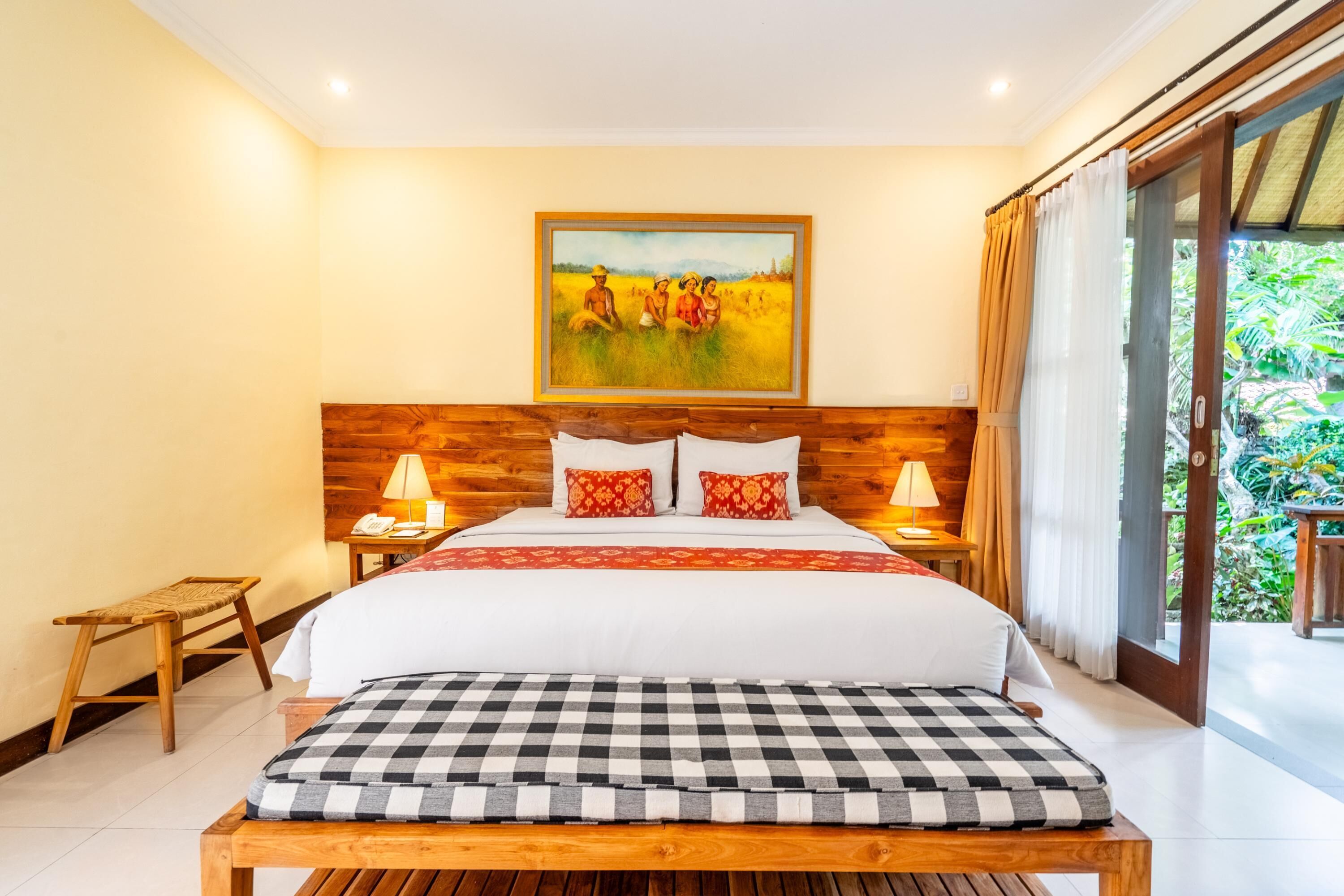Une chambre avec une tête de lit en bois, un grand lit avec une literie blanche et rouge, un banc à carreaux, une table de chevet avec une lampe et un tableau représentant un champ accroché au mur.