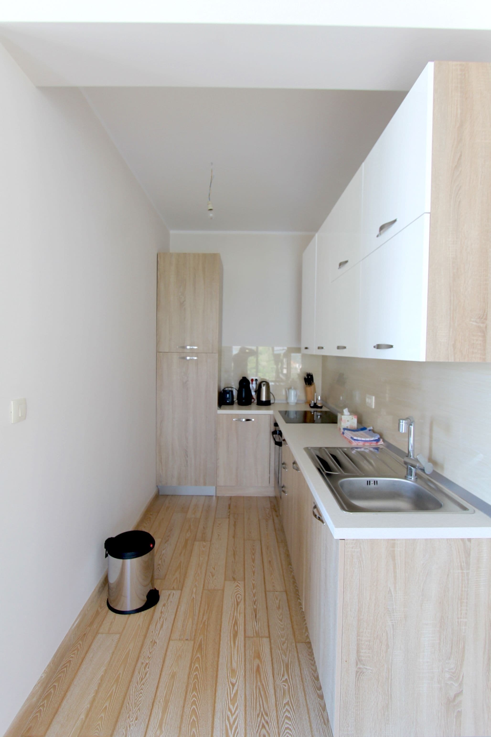 A compact kitchen with light wood cabinets, a stainless steel sink, and a small trash bin.