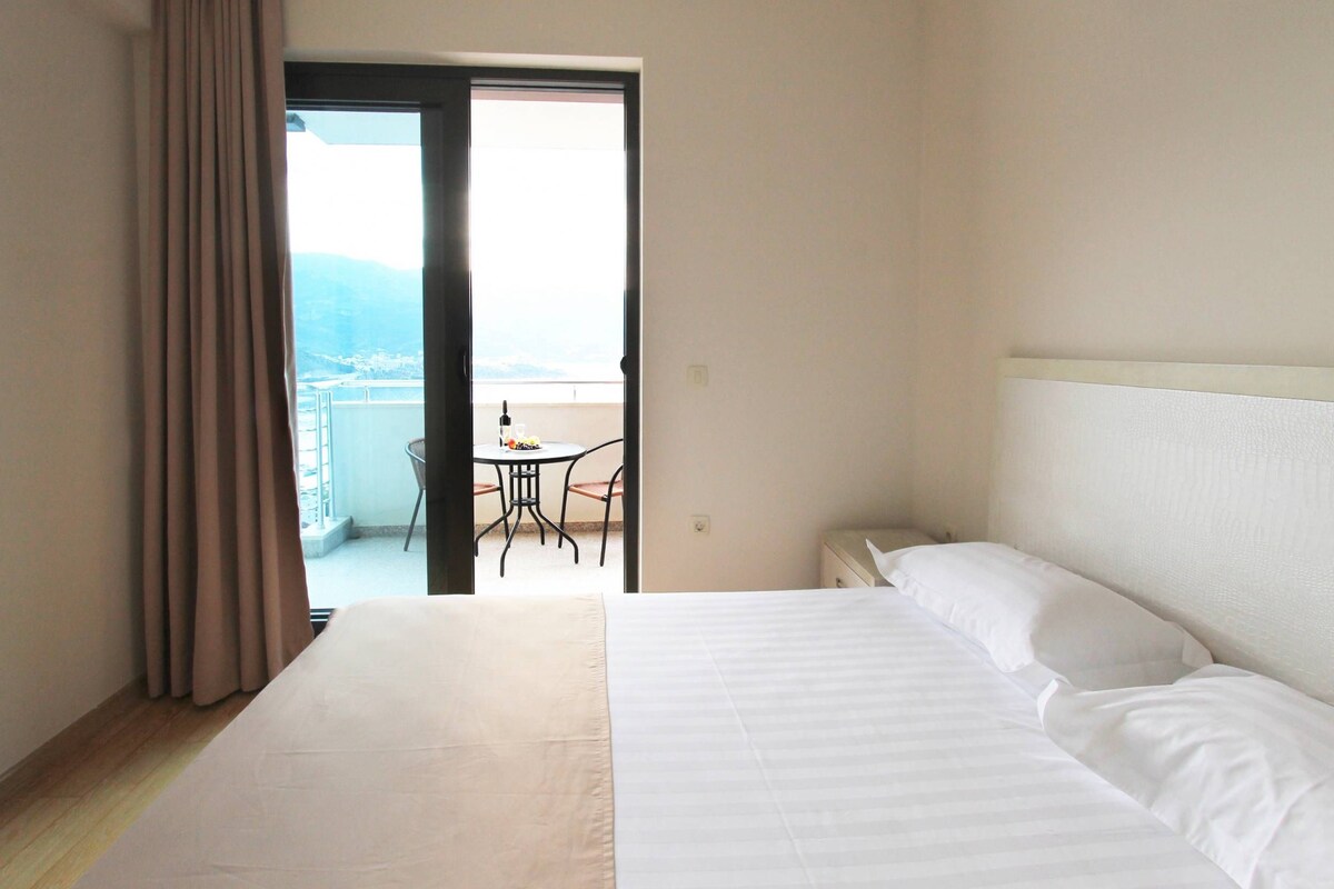 Une chambre d’hôtel avec un lit, une fenêtre avec des rideaux et un balcon avec une table et des chaises.