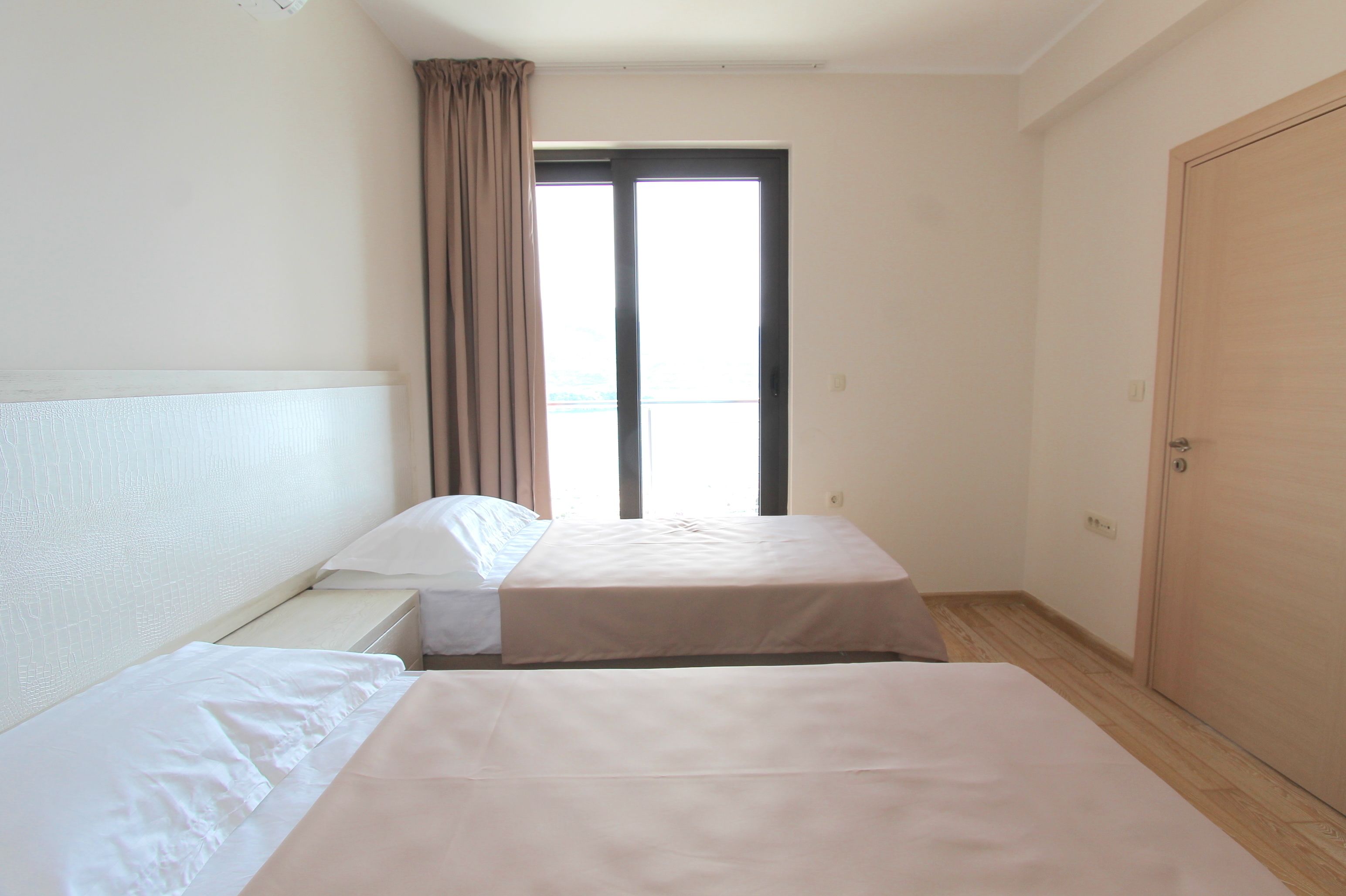 A hotel room with two beds, a window with curtains, a wooden door, and a view of the outside.