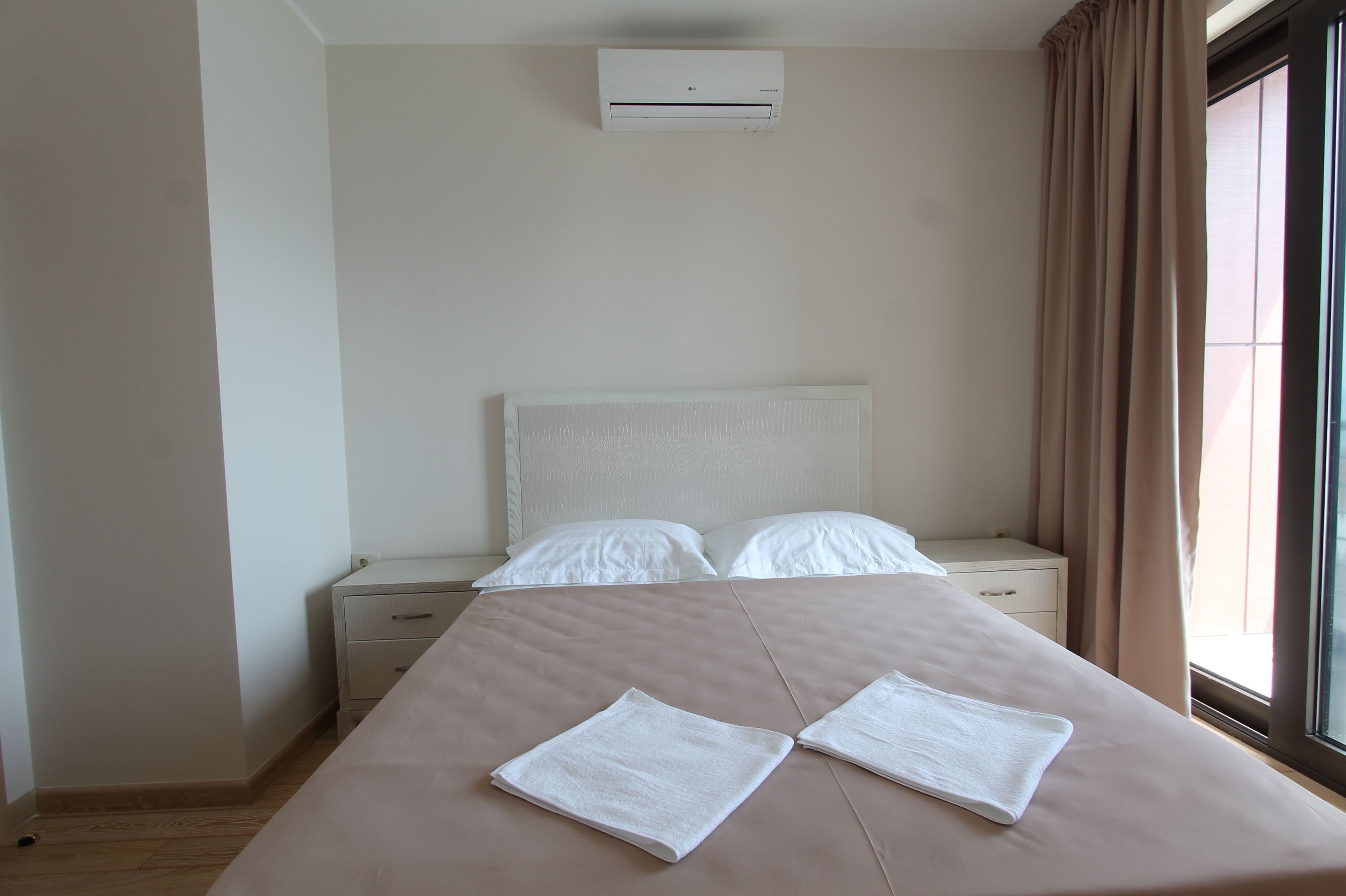 A neatly made bed with two folded towels, a white headboard, a bedside table with drawers, a window with curtains, and an air conditioning unit on the wall.