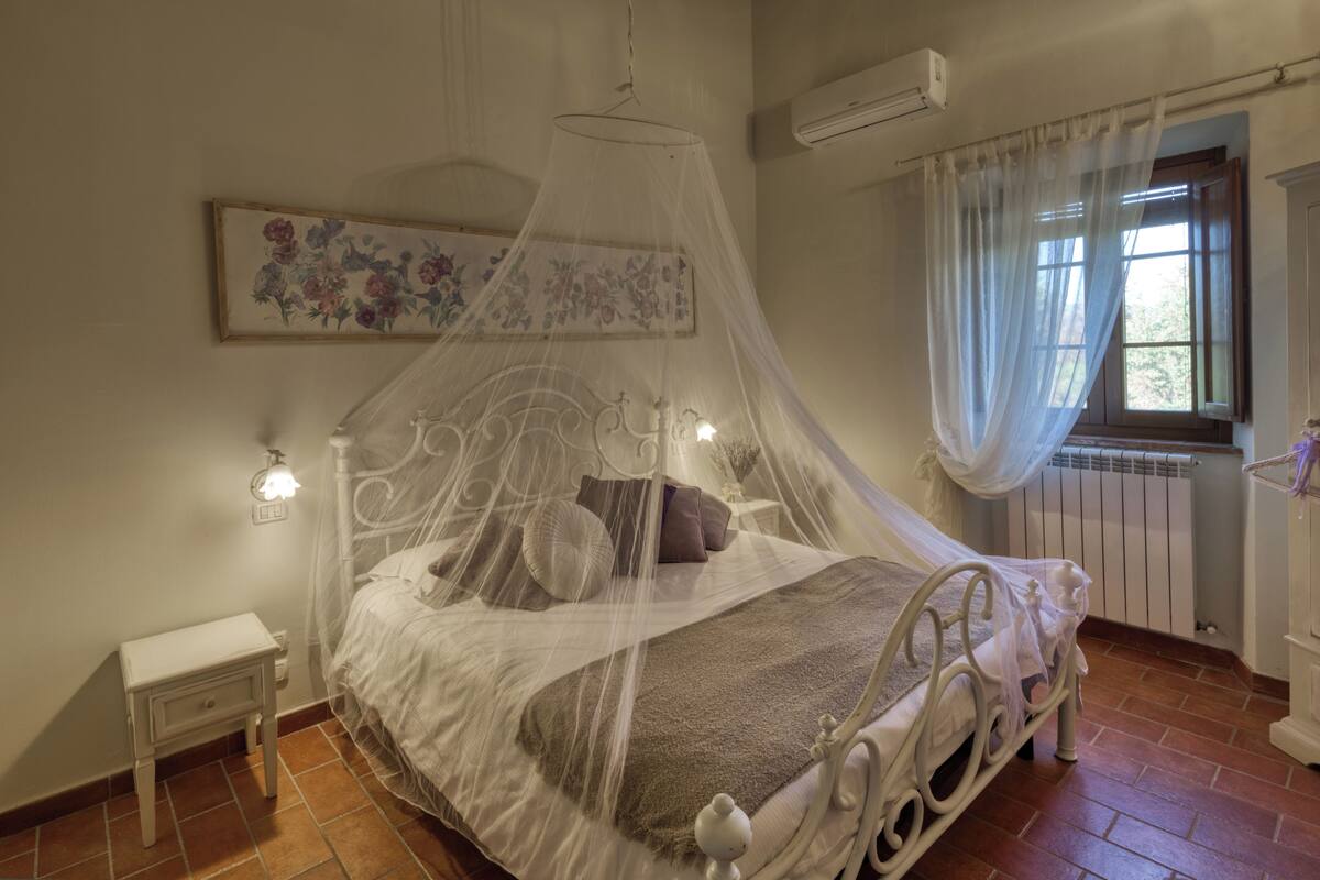Un dormitorio con cama con dosel, una mesita de noche, un armario de noche, una ventana con cortinas y un equipo de aire acondicionado.