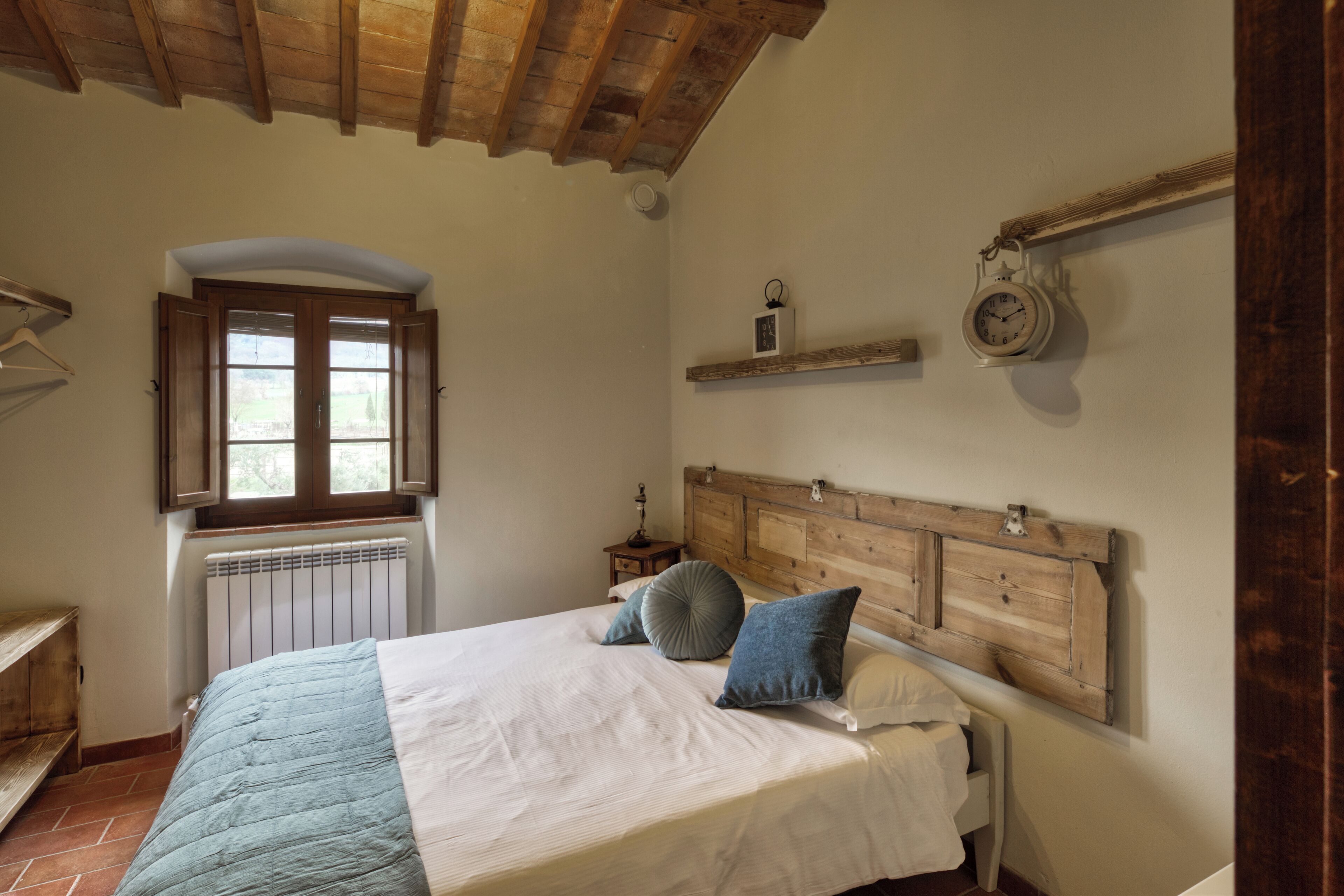 Un dormitorio con cabecera de madera, cama con edredón azul y blanco, ventana con persianas, radiador y estante con un reloj.