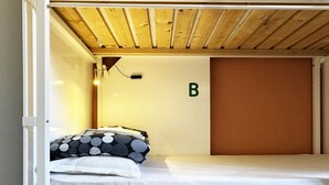 Ein Etagenbett mit einem Einzelbett oben und einem unterem Bett. Über dem Bett befindet sich eine wandmontierte Leuchte, und an der Wand ist ein Buchstabe 'B' zu sehen.