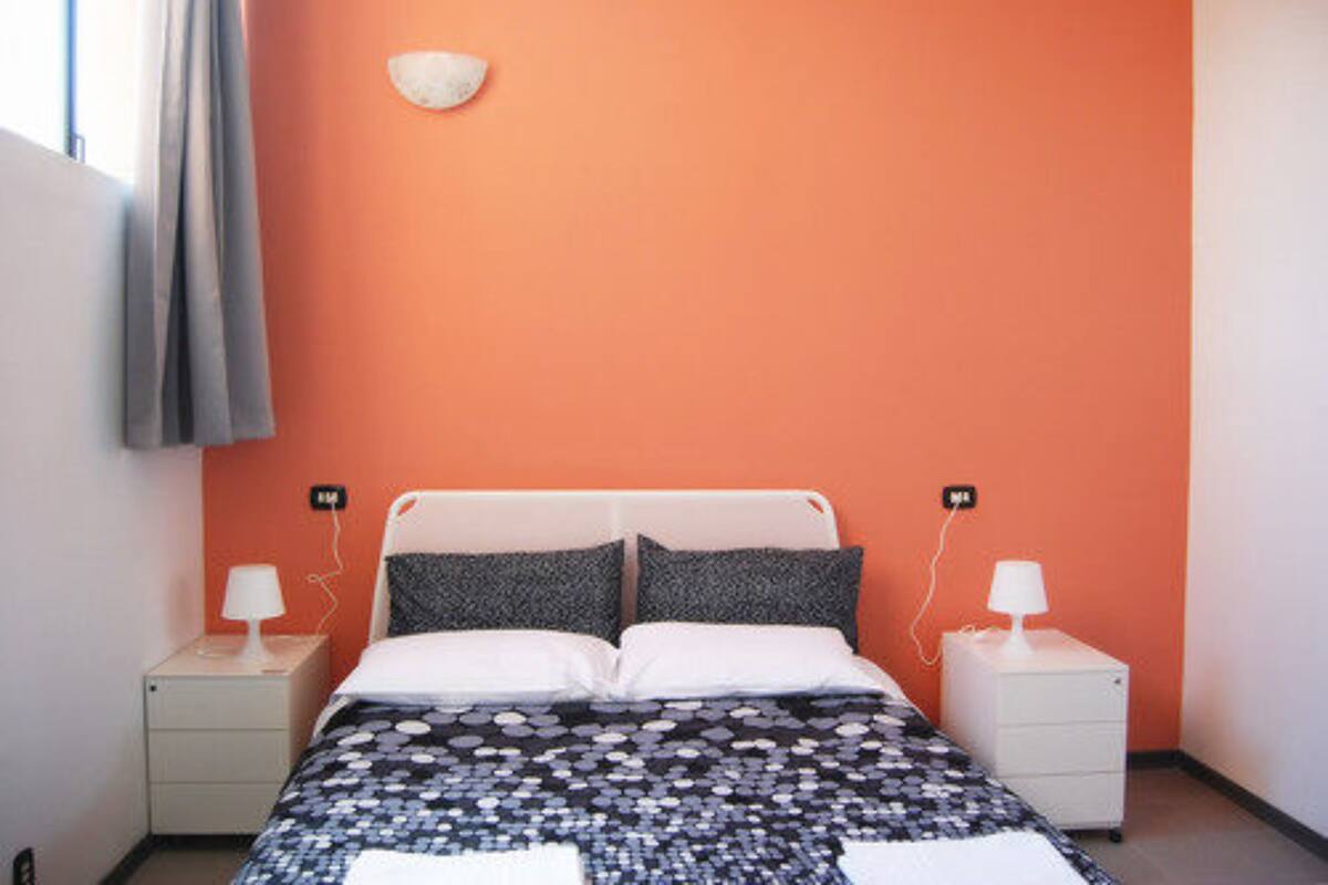 A bedroom with an orange wall, a white bed, two bedside tables with lamps, and a small wall-mounted light fixture.