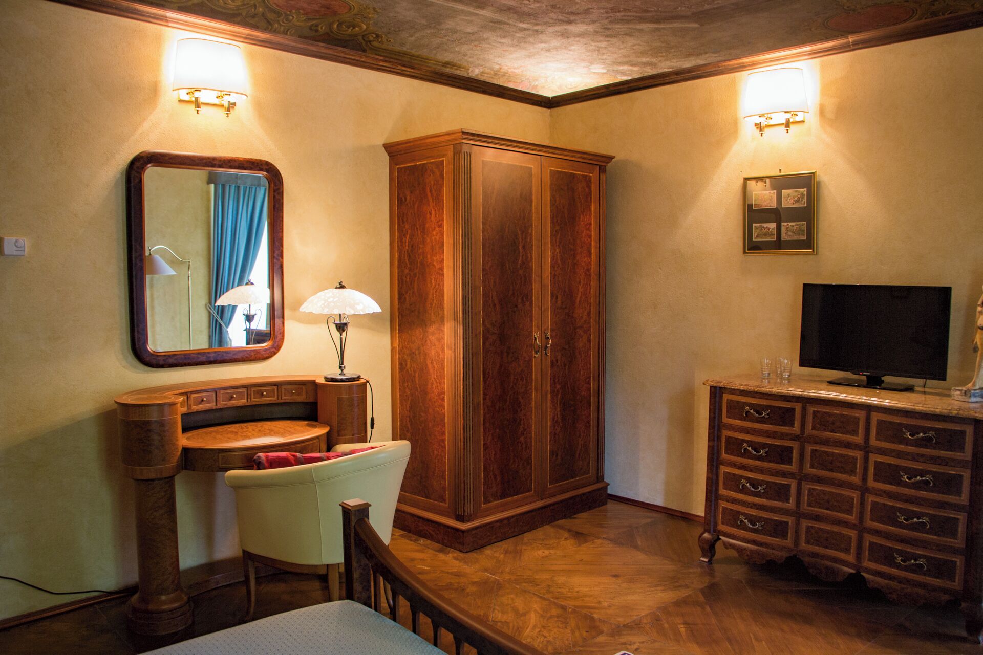 A well-lit room with a wooden wardrobe, a television on a cabinet, a desk with a chair, and a mirror with a blue curtain.